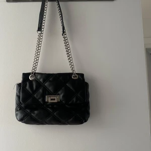 Svart quiltad axelväska med kedja - Snygg svart axelväska med quiltat mönster och kedjeaxelrem. Väskan har en elegant låsdetalj framtill och är perfekt för att ge en stilren touch till din outfit.