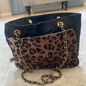 Leopardmönstrad axelväska med kedja - Snygg axelväska med leopardmönster och svarta detaljer. Väskan har en kedja som axelrem och en stilren design med små nitar på locket. Perfekt för att ge din outfit en trendig touch. Kom med bud! 