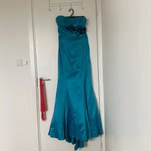 Elegant turkos långklänning i satin med axelbandslös design. Klänningen har vackra blommiga detaljer vid bysten och en figurnära passform som framhäver silhuetten. Perfekt för festliga tillfällen.
