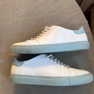 Arigato Skor - Snygga vita sneakers med en ljusblå sula och snörning. Perfekta för en stilren look. Märket är diskret tryckt på sidan av skorna.  Arigato Skor i bra skick