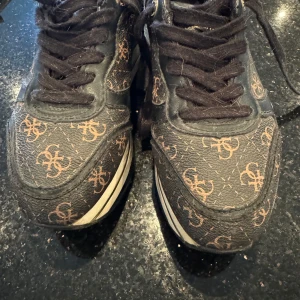 Svarta sneakers från Guess - Snygga svarta sneakers från Guess med brunt mönster och gulddetaljer. Skorna har snörning och en rund tå. Perfekta för en stilren look.