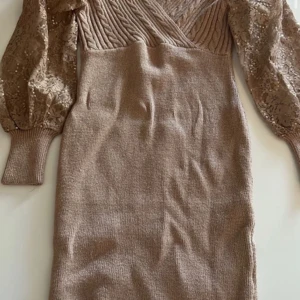 Beige stickad klänning med spetsärmar - Snygg beige stickad fest klänning med omlott-design och långa spetsärmar. Perfekt för en elegant look med en touch av romantik. Klänningen har en åtsittande passform och ribbade detaljer.