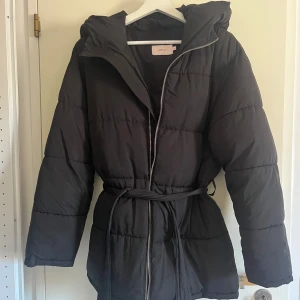 Svart dunjacka från Nelly - Säljer en svart dunjacka från Nelly med dragkedja och bälte i midjan. Jackan har långa ärmar och en mysig huva, perfekt för kyliga dagar.