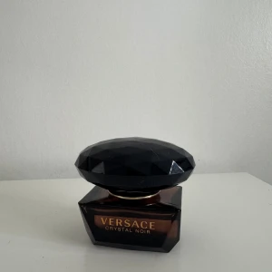 Versace Crystal Noir Parfym - Elegant parfymflaska från Versace med namnet Crystal Noir. Flaskan har en mörk, nästan svart färg och en unik, facetterad design som påminner om en ädelsten. Den lyxiga känslan förstärks av den guldiga texten på framsidan. Perfekt för den som söker en sofistikerad doftupplevelse.