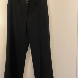 Svarta byxor från Lindex - Snygga svarta byxor från Lindex med tre knappar framtill. Byxorna har en rak passform och är perfekta för en stilren look. Aldrig använda!