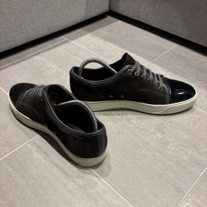 Bruna Lanvins  - Snygga svarta sneakers från Lanvins med glänsande lackdetaljer på captoe och snörning. Skorna har en vit sula som ger en fin kontrast till det svarta. Perfekta för en stilren look. Dustbag, box extra snören följer naturligtvis med. Uk8 men är stor i storlek. 42-43. Skick: 9.5/10