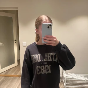 Mörkgrå sweatshirt från Gina Tricot - Jag säljer en mörkgrå/svart sweatshirt från Gina Tricot med ett tryck där fram 'ATHL. DEPT AMERICA 1983' i grå/beige text. Den kommer inte till användning så säljer den istället, pris kan diskuteras❤️