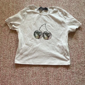 Vit t-shirt med discokörsbär från Primark - Säljer en vit t-shirt från Primark med ett coolt tryck av discokulor formade som körsbär. Perfekt för att ge din outfit en unik touch. T-shirten har korta ärmar och en rund halsringning.