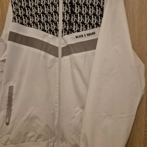 Vit vindjacka från Black X Squad - Säljer en stilren vit vindjacka från Black X Squad med svart mönster upptill och grå detaljer. Jackan har dragkedja framtill och två fickor med dragkedjor. Perfekt för en sportig look!