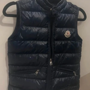 Svart dunväst från Moncler - Snygg svart dunväst från Moncler med dragkedja och två fickor framtill. Perfekt för kyligare dagar och ger en stilren look. Västen har en hög krage och Moncler-loggan på bröstet.