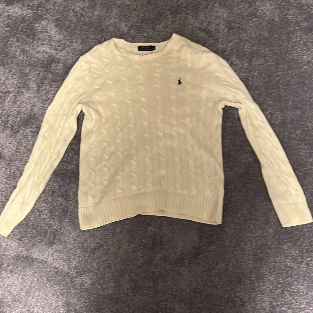 Säljer en stilren beige stickad tröja från Polo Ralph Lauren med kabelstickat mönster och den klassiska loggan på bröstet. Tröjan har långa ärmar och ribbade kanter för en snygg passform. Perfekt för kyligare dagar!. Neuletakit & Villapaidat.