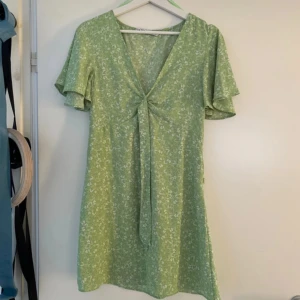 Grön blommig klänning - Säljer en söt grön klänning med blommigt mönster och korta ärmar. Klänningen har en v-ringad design med knytning framtill och volangdetaljer vid ärmarna.