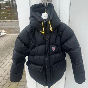Svart dunjacka från Fjällräven - Säljer en svart dunjacka från Fjällräven med huva och gula snören. Jackan har långa ärmar och en praktisk dragkedja med knappar framtill. Perfekt för kalla dagar.
