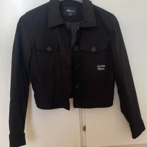 Svart tunn vårjacka från FB Sister - Snygg svart jacka från FB Sister med knappar framtill och två fickor på bröstet. Jackan har en croppad design och är perfekt för en stilren look. Märket [less] ISSUES pryder ena fickan. Den är perfekt att ha nu till våren och är lätt att stajla. Den är helt ny och oanvänd. Köpt från New Yorker 