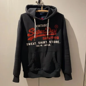 Svart hoodie från Superdry - Säljer en svart hoodie från Superdry med röd och vit text på framsidan. Den har en klassisk känguruficka och justerbar huva med vita snören. Perfekt för en avslappnad stil.