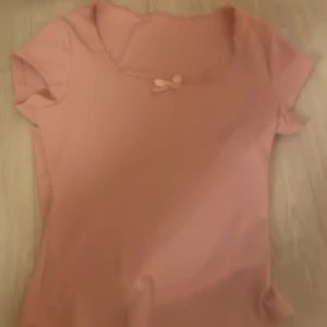 Rosa topp med spetsdetalj och rosett - Söt rosa topp med korta ärmar och en liten rosett framtill. Toppen har en fin spetsdetalj vid halsringningen som ger en romantisk touch. Perfekt för en feminin look! Provat 1 gång men det var inget för mig, inga hål/ fläckar!💕