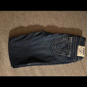 Mörkblå jeans från True Religion - Snygga mörkblå jeans från True Religion med låg midja och klassisk femficksdesign. Jeansen har en stilren look med kontrastsömmar och en ikonisk bakficka med broderad detalj. Perfekta för en trendig vardagsstil. Innerbens längd 76 cm