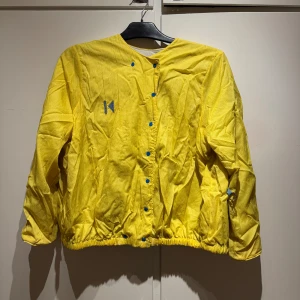 Gul jacka med blå knappar strl L uppskattningsvis - Säljer en cool gul jacka med blå knappar och en unik design. Jackan har långa ärmar och en elastisk nederkant. Perfekt för att lysa upp din outfit!