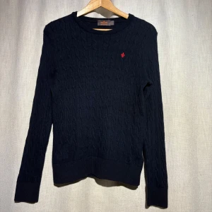 Mörkblå tröja från Morris - Säljer en stilren mörkblå tröja från Morris i 100% merinoull. Tröjan har ett diskret kabelstickat mönster och en liten röd broderad detalj på bröstet. Perfekt för kyligare dagar.