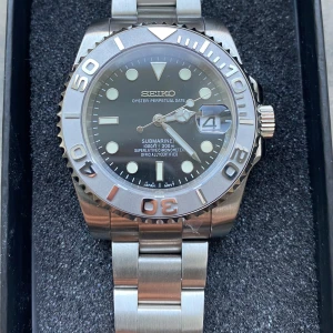 Silverfärgad klocka från Seiko - Snygg och robust klocka från Seiko med en silverfärgad boett och armband. Utrustad med en svart urtavla och tydliga markeringar. Klockan har en datumvisning och en vridbar bezel. Perfekt för den som gillar en klassisk och stilren design.