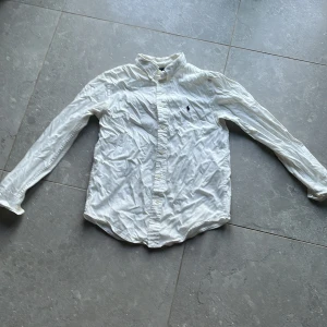 Vit skjorta från Ralph Lauren - Säljer en klassisk vit skjorta från Ralph Lauren med broderad logga på bröstet. Skjortan har långa ärmar och knappar framtill. Perfekt för en stilren look. Obs! Liten fläck se sista bilden!