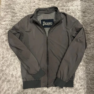 Grå jacka från Herno - Snygg grå jacka från Herno med dragkedja framtill. Jackan har långa ärmar och ribbade muddar vid ärmslut och nederkant. Perfekt för en stilren look.