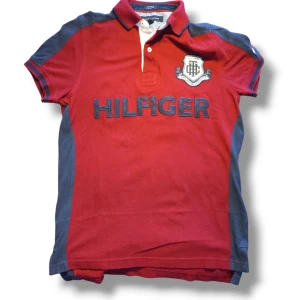 Röd pikétröja från Tommy Hilfiger - Skit fet polo. Säljer en snygg röd pikétröja från Tommy Hilfiger med blå detaljer och ett stort Hilfiger-tryck på framsidan. Tröjan har korta ärmar och en klassisk krage med knappar. Perfekt för en stilren look.