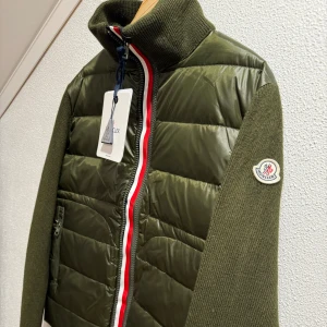 Olivgrön dunjacka från Moncler - Snygg olivgrön dunjacka från Moncler med stickade ärmar och quiltad front. Jackan har en dragkedja med röd och vit kant, samt Moncler-logga på ärmen. Perfekt för kyliga dagar med stil.