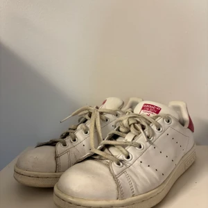  Stan Smith sneakers - Säljer ett par jättefina klassiska vita Adidas Stan Smith sneakers med rosa detaljer på hälen. Skorna har snörning och är perfekta för en stilren och sportig look. Storlek 38. 