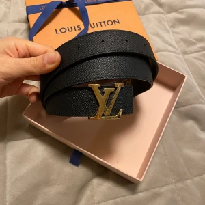 Svart och brunt bälte från Louis Vuitton - Snyggt bälte från Louis Vuitton med en ikonisk LV-spänne i guld. Ena sidan är svart och den andra har det klassiska bruna monogrammönstret. Perfekt för att ge din outfit en lyxig touch.