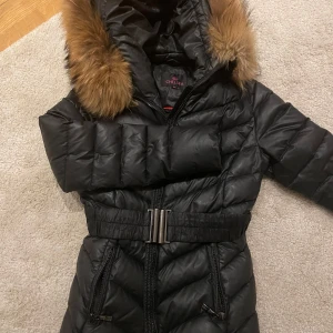 Svart dunjacka från Chelsea - Snygg svart dunjacka från Chelsea med pälskantad huva och bälte i midjan. Jackan har dragkedja framtill och är perfekt för kalla dagar. Den quiltade designen ger en stilren look.