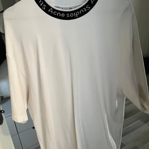 Vit t-shirt från Acne Studios - Säljer en stilren vit t-shirt från Acne Studios med en svart rundad krage där märkesnamnet är tryckt. T-shirten är tillverkad i en mjuk blandning av viskos och nylon. Storleken är xs men sitter mer som M 