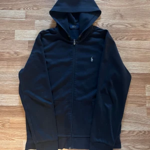 Svart hoodie från Polo Ralph Lauren - Svart hoodie från Polo Ralph Lauren med dragkedja framtill och klassisk huva. Tröjan har det ikoniska broderade logotypen på bröstet och två fickor framtill. Perfekt för en chill och stilren look.