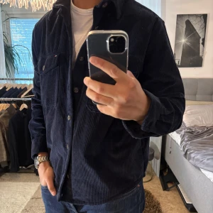 Mörkblå manchester overshirt Tommy Hilfiger - Snygg mörkblå overshirt från Tommy Hilfiger i manchester. Avslappnad passform med två bröstfickor med knappar och klassisk krage. Perfekt lager-på-lager-plagg med tydliga Hilfiger-detaljer och knäppning framtill.