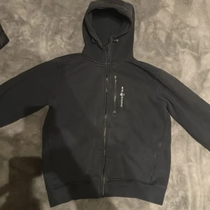 Svart hoodie från Sail Racing - Svart hoodie från Sail Racing med hel dragkedja och bröstficka med dragkedja. Snygg logga på bröstet och justerbar huva. Perfekt för dig som gillar stilren och sportig look. Materialet är mjukt och känns bekvämt mot huden.