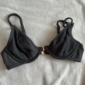 Svart bikiniöverdel från Lounge underwear - Svart bikiniöverdel från Lounge med snygg vågig struktur i tyget och guldfärgade metalldetaljer. Justerbara axelband och smidig knäppning framtill. Använd 1 gång superbra skick.