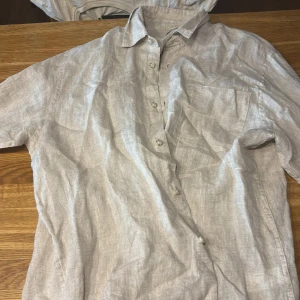 Beige linneskjorta från Kappahl M - Luftig och stilren beige skjorta från Kappahl i storlek M. Skjortan är tillverkad i linne, har korta ärmar, klassisk krage, bröstficka och knäppning framtill. Perfekt för varma dagar när du vill ha en avslappnad men snygg look.