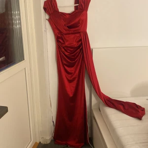Röd långklänning i satin - Snygg röd långklänning med glansig satin-känsla och draperad detalj framtill. Klänningen har breda axelband, markerad midja och en lång släp som ger extra drama. Perfekt för bal eller fest där du vill sticka ut. Använd 1 gång