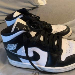 Nike Air Jordan 1 svartvita sneakers - Snygga Nike Air Jordan 1 sneakers i svart och vitt skinn med klassisk high-top design. Skorna har svart snörning, perforerad tå och ikoniska Jordan-loggan på sidan. Perfekta för dig som vill ha en stilren och sportig look.