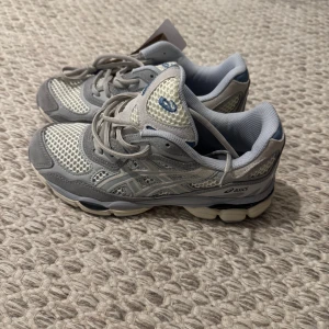 Asics gel nyc - Snygga Asics sneakers i grått och vitt med meshpaneler och silvriga detaljer. Skorna har snörning, rund tå och en chunky sula i beige och grå. Perfekta för dig som gillar sportig stil och komfort. Klassisk Asics-logga på sidan.
