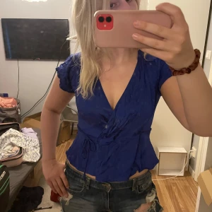 Blå omlottopp med knyt i midjan - Snygg blå omlottopp med korta ärmar och djup v-ringning. Toppen har knyt i midjan som ger en feminin siluett och lätt volang nertill. Perfekt att styla med jeans eller kjol för en trendig look.