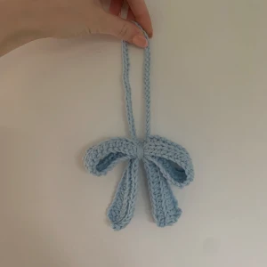 Handvirkad ljusblå rosett-accessoar - Supersöt handvirkad rosett i ljusblått garn, perfekt att använda som accessoar till väskor och nyckelringar. Rosetten är mjuk och har en enkel ögla för att enkelt kunna fästas där du vill. Ger en gullig och personlig touch till din stil.