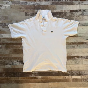 Vit Lacoste pikétröja  - Passar size S. Klassisk vit pikétröja från Lacoste med korta ärmar och ikoniska krokodilloggan på bröstet. Tröjan har krage och två knappar framtill. Tillverkad i mjuk bomull som andas och passar perfekt till en avslappnad stil. 
