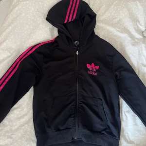 Svart Adidas hoodie med rosa detaljer - Svart hoodie från Adidas med dragkedja, rosa Adidas-tryck och ikoniska rosa ränder längs ärmar och huva. Stor logga på ryggen och mindre framtill. Perfekt för dig som gillar sportig och street-inspirerad stil. Liten i storleken!