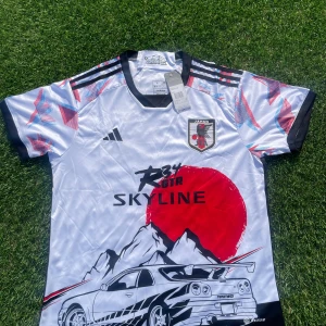 Japan Skyline x Adidas fotbollströja L - Unik Japan fotbollströja från Adidas i storlek L, med coolt Skyline R34 GTR-tryck, stora röda solskivan, berg och Nissan-detaljer. Tröjan är vit med svarta och röda inslag, korta ärmar och grafiskt mönster på axlarna. Tillverkad i lätt och ventilerande material.