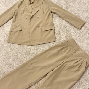 Beige kostym med kavaj och byxa - Elegant set bestående av en kavaj och matchande byxor. Kavajen har en dubbelknäppning och dekorativa fickor framtill. Byxorna har en klassisk, rak passform.   Byxornas längd : 99,5 Midja: 61 Överdelens längd : 65,5 Byst : 104  Axel : 37