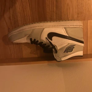 Nike Air Jordan 1 High vit/svart - Snygga Nike Air Jordan 1 High sneakers i vitt och grått skinn med svarta detaljer och klassisk svart swoosh. Skorna har svarta snören, perforerad tå och ikonisk Air Jordan-logga på sidan. Perfekta för dig som vill sticka ut med en tidlös streetstil.