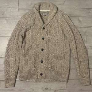Beige stickad kofta från Next - Säljer en beige stickad kofta från Next med storlek S. Den  har grovt kabelstickat mönster, sjalkrage och stora knappar framtill. Koftan har två praktiska fickor och ribbade muddar. Perfekt för lager-på-lager och chill höst- och vinterstil. Varan är använd Max 7 tillfällen och nypris ligger den på 800 kr