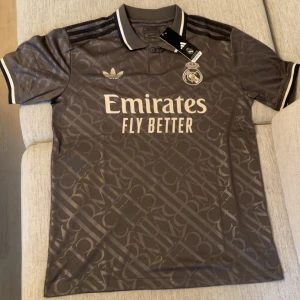 Real Madrid x Adidas grå fotbollströja - Snygg brun Real Madrid fotbollströja från Adidas med diskret mönster över hela tyget. Tröjan har korta ärmar, klassisk krage och svarta Adidas-ränder på axlarna. Broderad Real Madrid-logga och Emirates Fly Better-tryck på bröstet. Perfekt för dig som älskar fotboll och streetstyle.