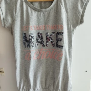 Grå t-shirt med paljetter och text - Snygg grå topp med korta ärmar och texten 'LET'S TAKE TIME TO MAKE a choice' framtill. Ordet MAKE är dekorerat med glittriga paljetter i svart och silver. Mjuk bomullskänsla och avslappnad passform, perfekt för en chill look.